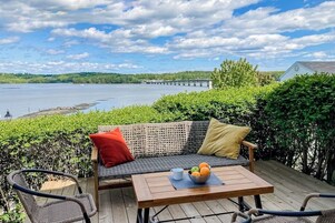 A deck with a wicker sofa, a table with fruit, and a view of a bridge over water.