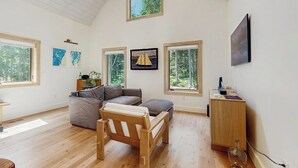 A spacious room with a high ceiling, large windows, a wooden chair, a sofa, and a small cabinet. - Charming New Home With Hot Tub, Spacious Deck, Gas Grill, Wifi, and Washer/dryer (East Boothbay)