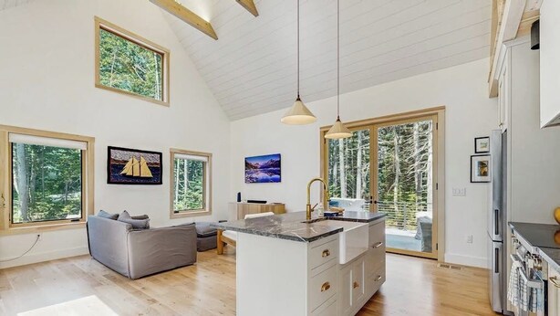 A modern kitchen with a central island, wooden flooring, and large windows offering a view of trees. - Charming New Home With Hot Tub, Spacious Deck, Gas Grill, Wifi, and Washer/dryer (East Boothbay)