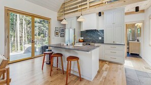A modern kitchen with a large island, bar stools, and a view of the outdoors. - Charming New Home With Hot Tub, Spacious Deck, Gas Grill, Wifi, and Washer/dryer (East Boothbay)
