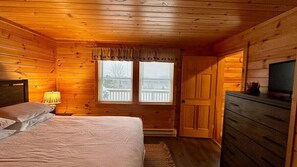 A wooden cabin bedroom with a bed, a dresser, a television, and a window with blinds. - Moosehead State of Mind, 3BR Cabin With Mtn Views & Deck (Shirley)