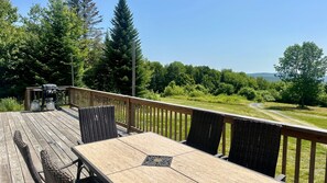 A wooden deck with wicker furniture and a grill, overlooking a grassy area and trees. - Moosehead State of Mind, 3BR Cabin With Mtn Views & Deck (Shirley)