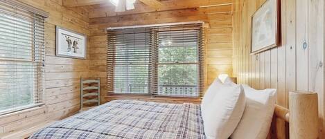 Ein Schlafzimmer einer Holzhütte mit einem Bett, einem Deckenventilator und zwei Fenstern mit Jalousien.