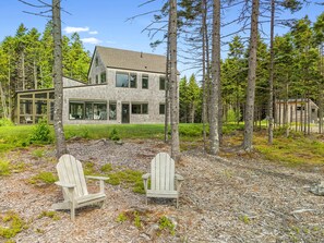 A modern two-story house surrounded by a forest, with two Adirondack chairs in the foreground. - Quiet, Modern, Dog-friendly Home With Ocean Views, Private Trails & Firepit (Tenants Harbor)