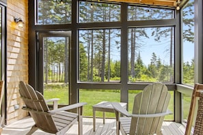 A wooden deck with two Adirondack chairs and a small table, overlooking a forested area through large windows. - Quiet, Modern, Dog-friendly Home With Ocean Views, Private Trails & Firepit (Tenants Harbor)