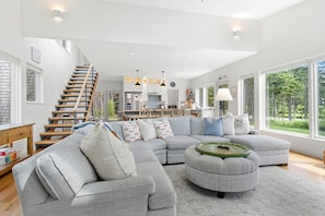A spacious living area with a large sectional sofa, a round ottoman, and a staircase leading to an upper level. - Quiet, Modern, Dog-friendly Home With Ocean Views, Private Trails & Firepit (Tenants Harbor)