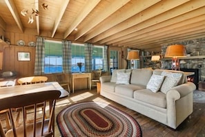 A cozy cabin interior with a wooden ceiling, a sofa, a dining table, and a fireplace. - Marshall Point Cottage, Classic 5-bedroom (Tenants Harbor)