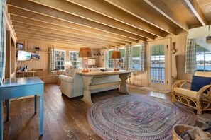 A spacious room with a wooden ceiling, a large window, a round woven rug, and a dining table. - Marshall Point Cottage, Classic 5-bedroom (Tenants Harbor)