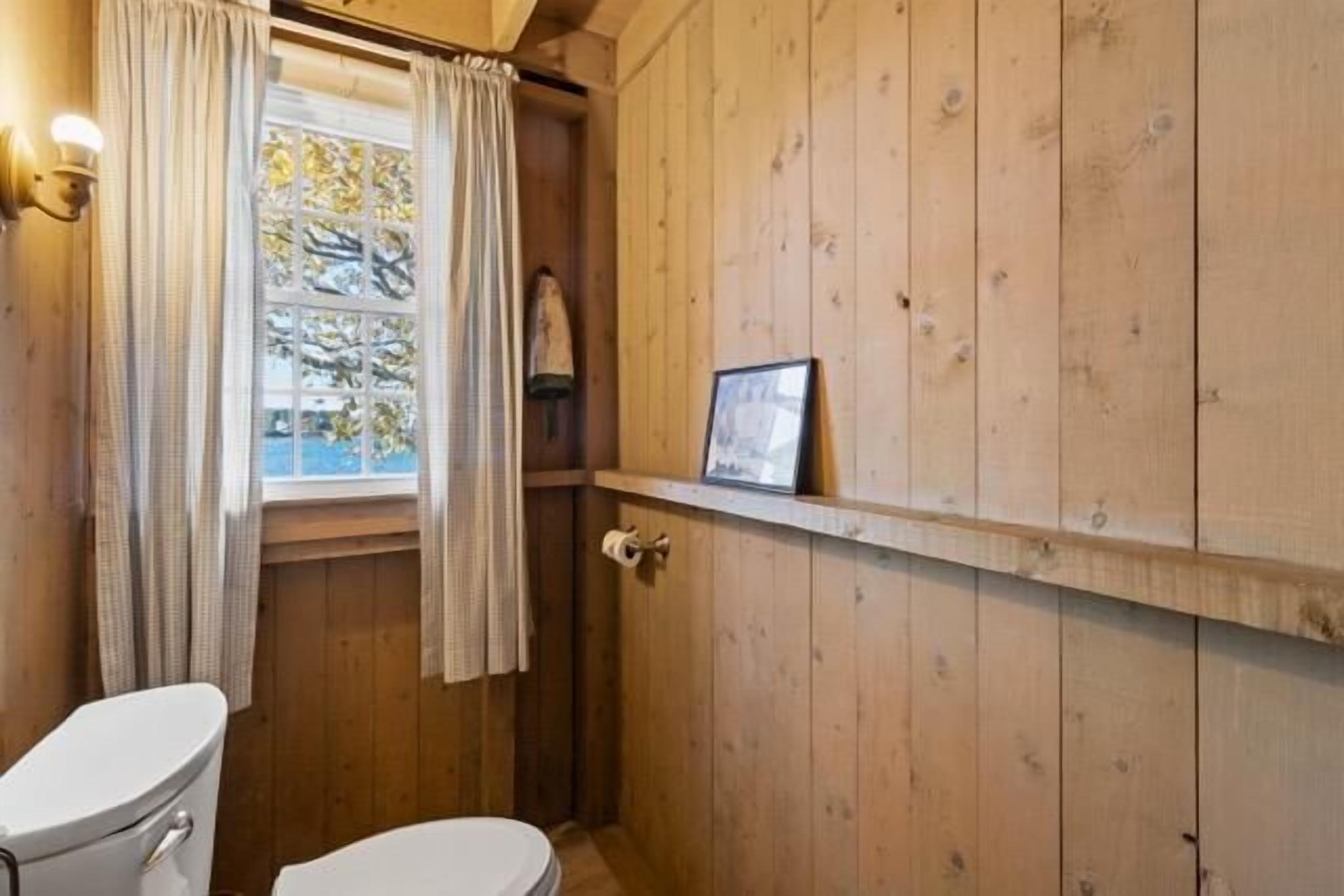 A wooden-paneled bathroom with a toilet, a window with curtains, and a framed picture on the wall.