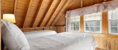 Un dormitorio de ático con suelo de madera, una cama individual apilada, una ventana con cortinas y una lámpara a la izquierda.