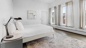 A bedroom with a bed, a bedside table with a lamp, a door, and a window with curtains. - Rockland Central (Rockland)