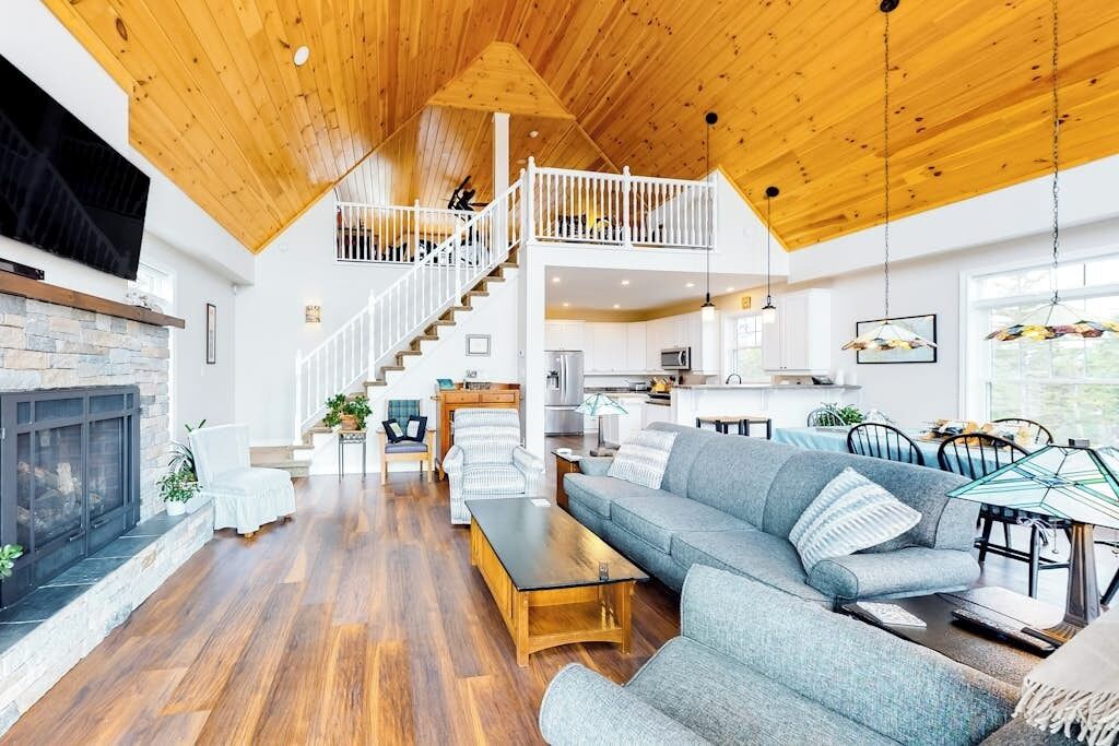 A spacious living area with a wooden ceiling, a stone fireplace, a grey sofa, and a dining area with a table and chairs.