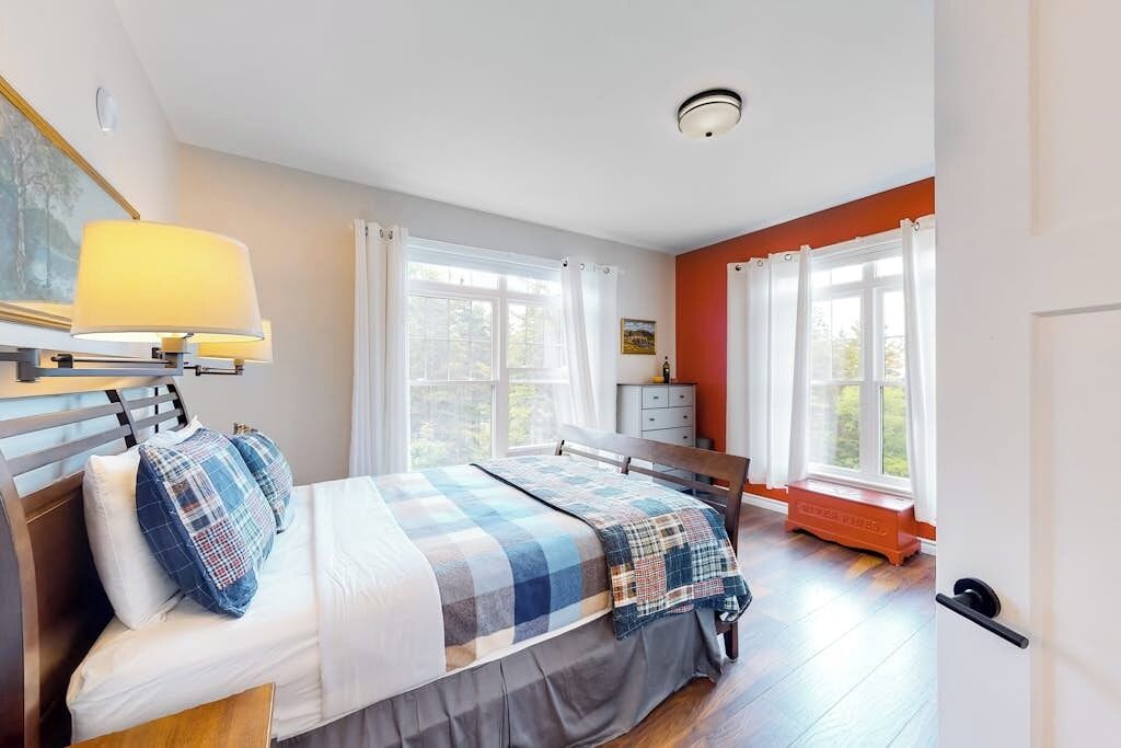 A bedroom with a bed, bedside table, lamp, windows, and a view of the outdoors.