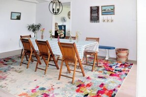 A dining area with a table set for four, surrounded by wooden chairs, a colorful rug, and a hanging light fixture. - Ocean-view Cottage Near Beach With Deck, Grill, & Full Kitchen (New Harbor)