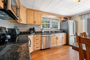 A kitchen with wooden cabinets, a stainless steel dishwasher, a black countertop, and a window with a view of trees. - Boothbay Harbor Home With Water Views Near Dining & Whale Watching (East Boothbay)