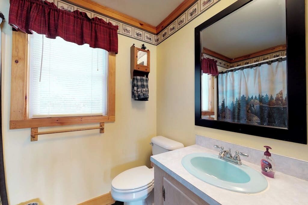 A bathroom with a toilet, sink, mirror, window with curtains, and a towel rack.