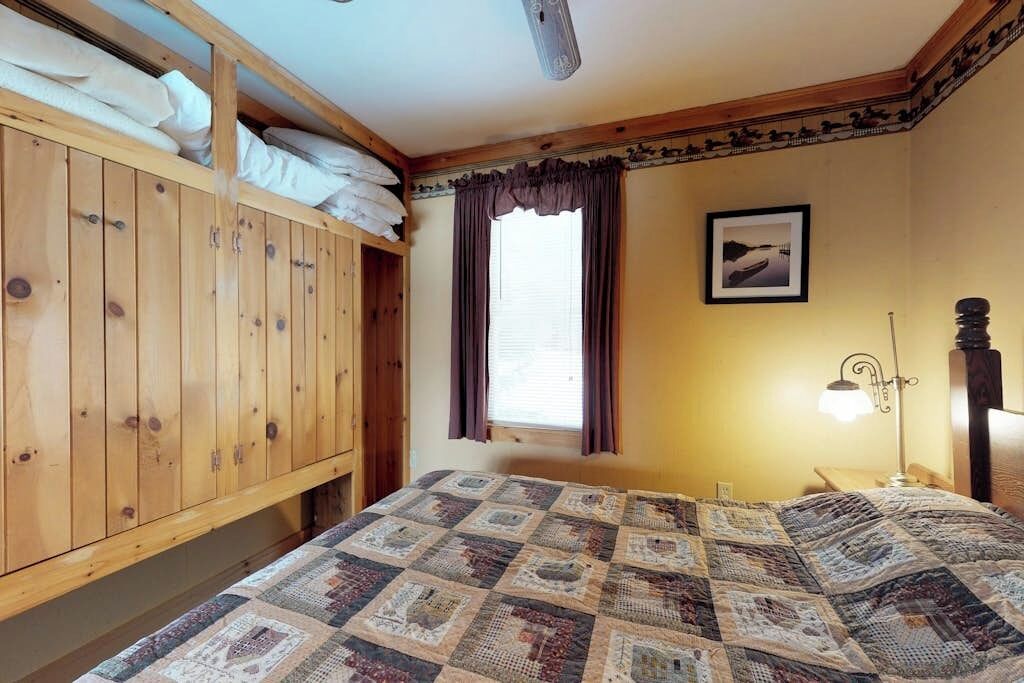 Una habitación con literas, una colcha acolchada, una ventana con cortinas, un cuadro enmarcado en la pared y una lámpara de noche.