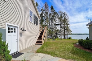 A house with a wooden staircase leading to a deck, surrounded by a grassy yard and tall trees. - Newly-built Waterfront Home With Fast Wifi, Furnished Deck, & Washer/dryer (Waldoboro)