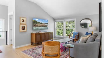 A living room with a high ceiling, a large window, a grey sofa, a wooden chair, a small table, and a television.