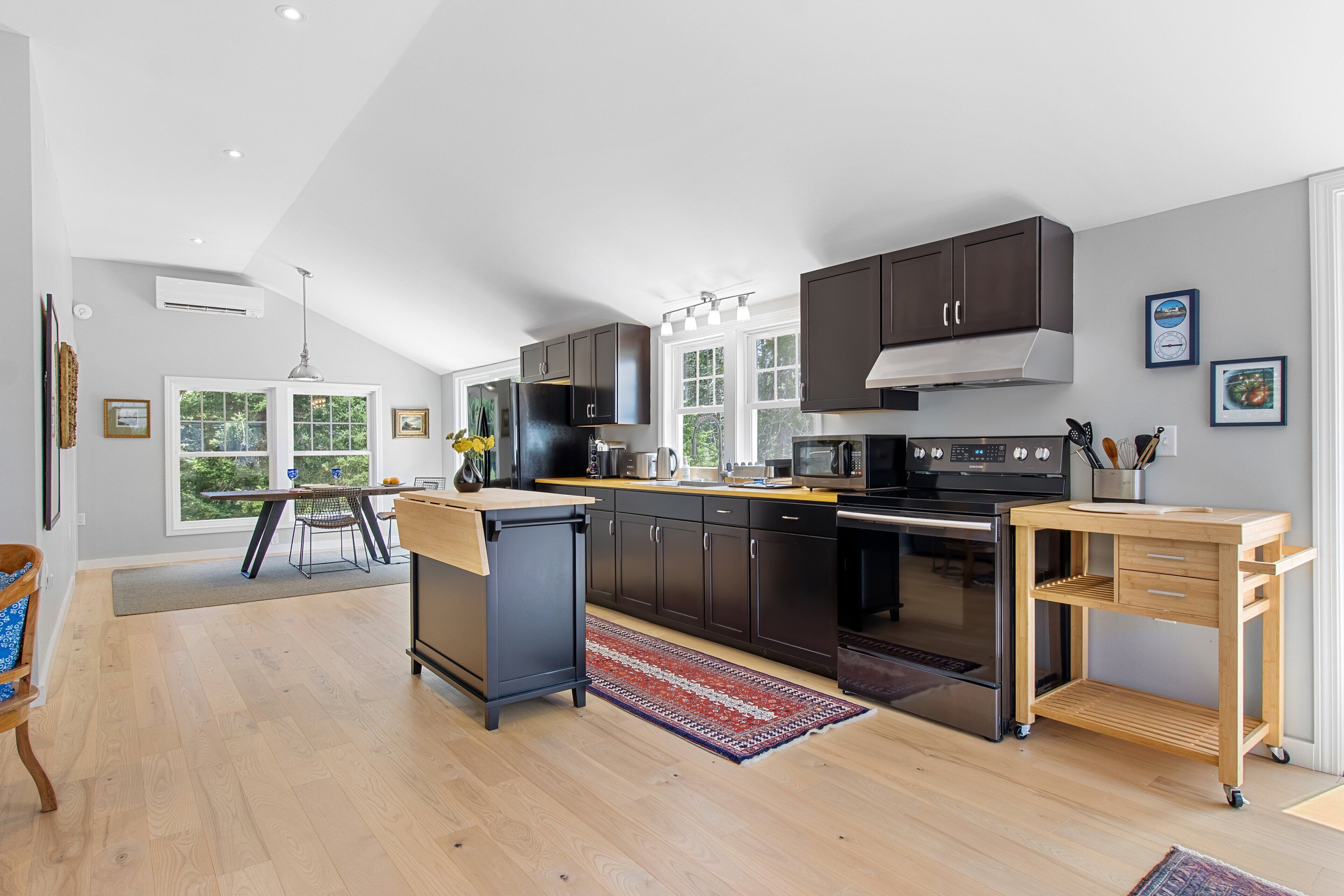 A modern kitchen with dark cabinetry, a wooden dining table, and a view of the outdoors through large windows.