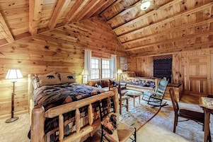 Interior de uma cabana de tronco com uma cama, cadeiras, uma mesa e uma janela.