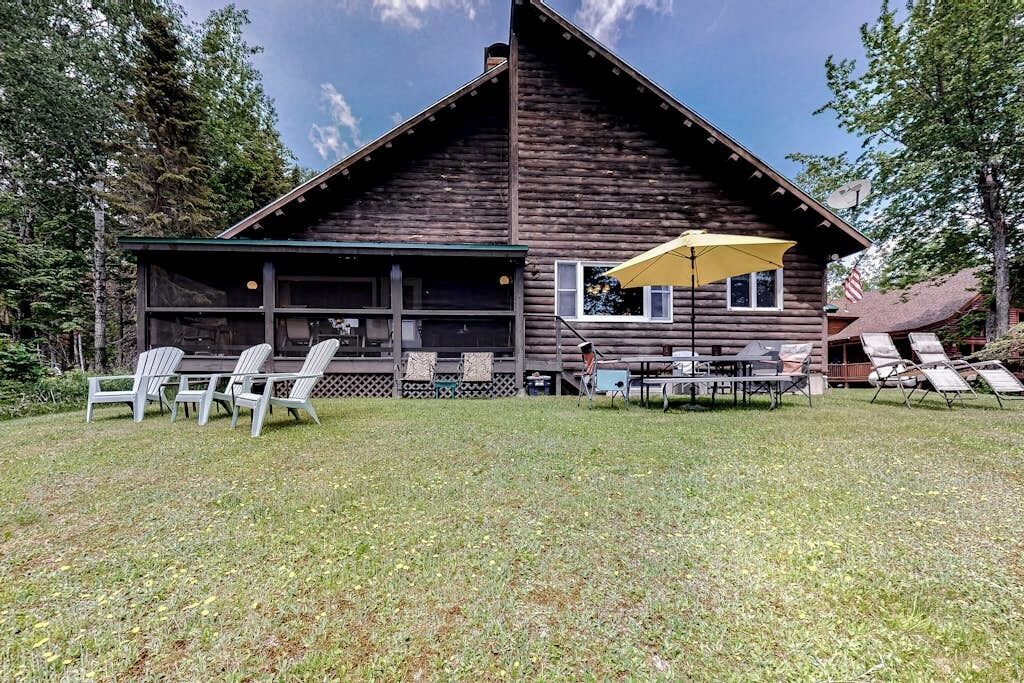 A rustic log cabin with a covered patio, outdoor seating, and a view of the surrounding forest.