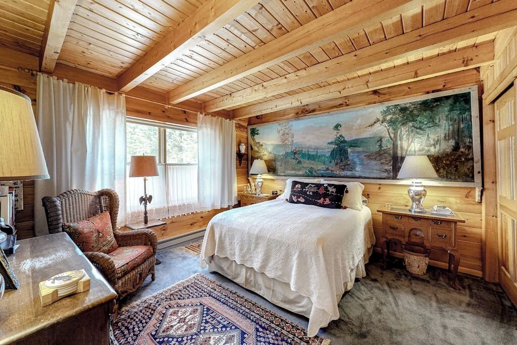 A wooden cabin bedroom with a bed, a chair, a desk, and a painting on the wall.