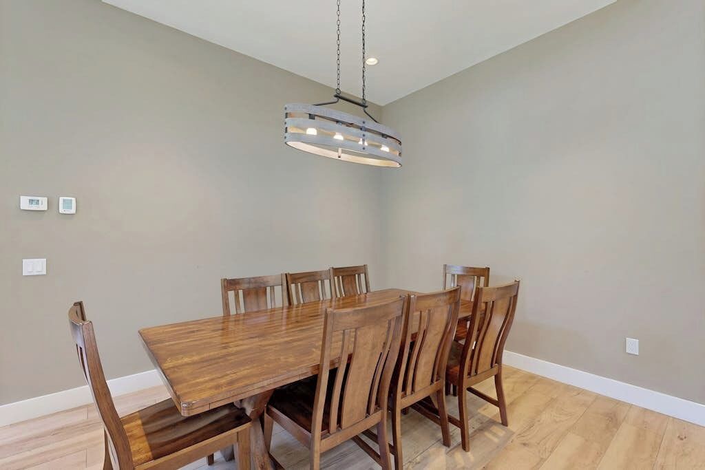 A dining area with a wooden table and chairs, a hanging light fixture, and a plain wall.