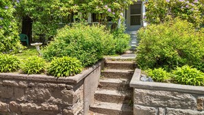 En stentrappa leder upp till ett hus med en veranda, omgivet av frodig grönska och blommande buskar.