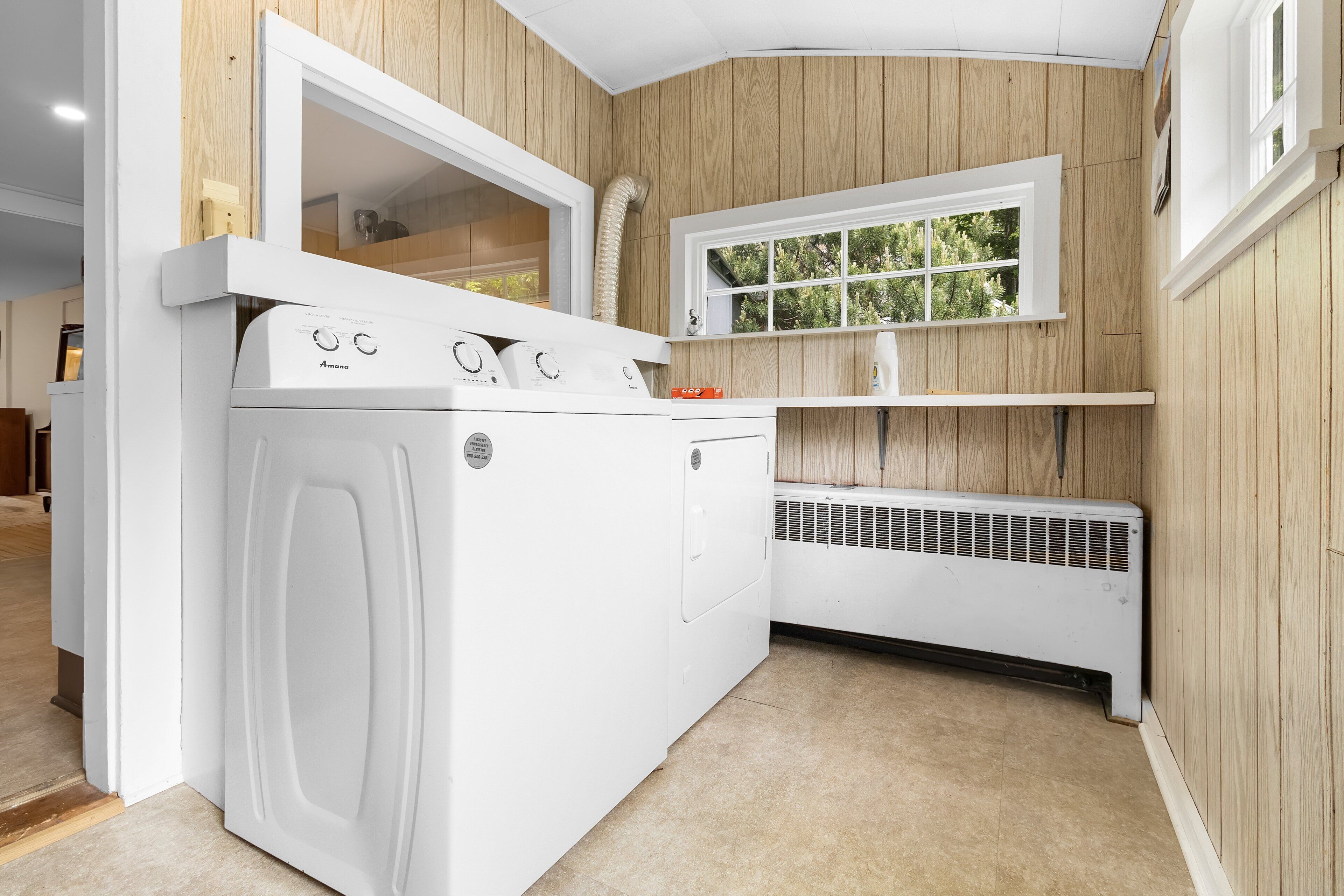 A laundry room with a white washer and dryer, a window with a view of trees, and wooden paneling on the walls.