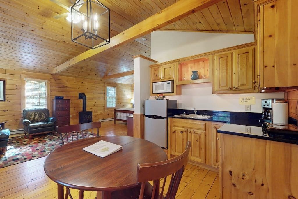 A wooden cabin interior with a dining table, kitchen area, and a fireplace.