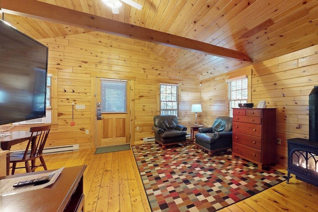A log cabin interior with a television, leather chairs, a fireplace, and a patterned rug.