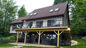 A two-story house with a covered patio, outdoor seating, and a stone wall. - Spacious Lakefront Home With a Sandy Beach, Kayaks, and Stunning Panoramic Views (Holden)