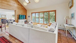 A spacious living room with a large sofa, a dining area, and a view of the outdoors. - Spacious Lakefront Home With a Sandy Beach, Kayaks, and Stunning Panoramic Views (Holden)