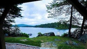 A view from a covered area overlooking a lake with a wooden deck, a stone pathway, and surrounding greenery. - Spacious Lakefront Home With a Sandy Beach, Kayaks, and Stunning Panoramic Views (Holden)