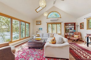 A spacious living room with large windows, a fireplace, a bookshelf, and a comfortable seating area. - Spacious Lakefront Home With a Sandy Beach, Kayaks, and Stunning Panoramic Views (Holden)