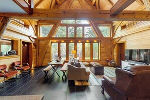 Interior de una cabaña de madera con un dormitorio en el altillo, una sala de estar con sofá y sillones, y una mesa de comedor.
