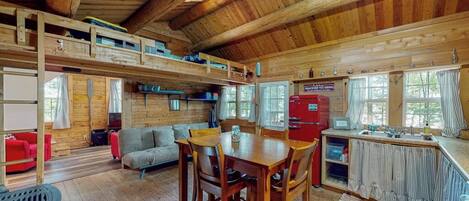 A wooden cabin interior with a dining area, kitchen, and upper sleeping quarters.