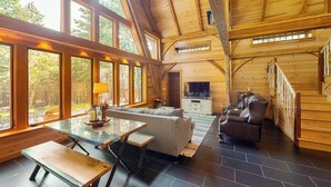 A spacious wooden cabin with large windows, a dining area, a living room with a sofa and a TV, and a staircase.