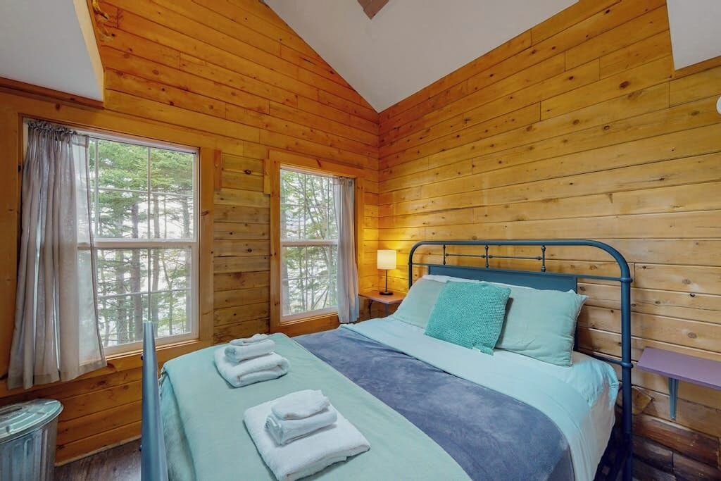 Quarto de cabana de madeira com cama, duas janelas, mesa de cabeceira e abajur.