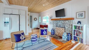 A modern living room with a stone fireplace, a television mounted above it, and a bookshelf filled with books. - Bayfront Cottage on Mill Cove w/ Private Dock & Furnished Deck - Walk Downtown (Boothbay Harbor)