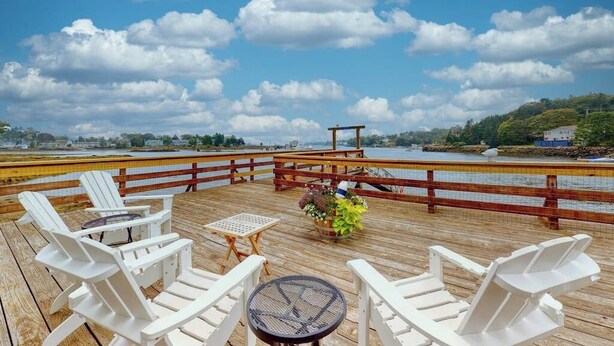 A wooden deck with white chairs and a small table, overlooking a body of water and a distant shoreline. - Bayfront Cottage on Mill Cove w/ Private Dock & Furnished Deck - Walk Downtown (Boothbay Harbor)
