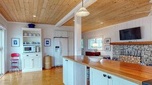 A modern kitchen with wooden ceiling, white cabinetry, a stone fireplace, and a TV mounted on the wall. - Bayfront Cottage on Mill Cove w/ Private Dock & Furnished Deck - Walk Downtown (Boothbay Harbor)