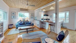 A spacious living area with a wooden ceiling, a kitchen with bar stools, and large windows offering views. - Bayfront Cottage on Mill Cove w/ Private Dock & Furnished Deck - Walk Downtown (Boothbay Harbor)
