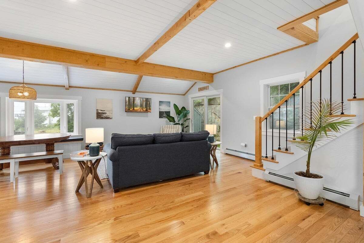 Een ruime woonkamer met een houten vloer, een donkere bank, een houten bankje, een kleine tafel met een lamp, en een trap met een plant in een pot.