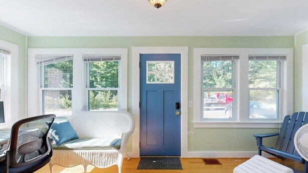 Una habitación con una puerta azul, muebles de mimbre blanco, un ventilador de pie y vistas al exterior.