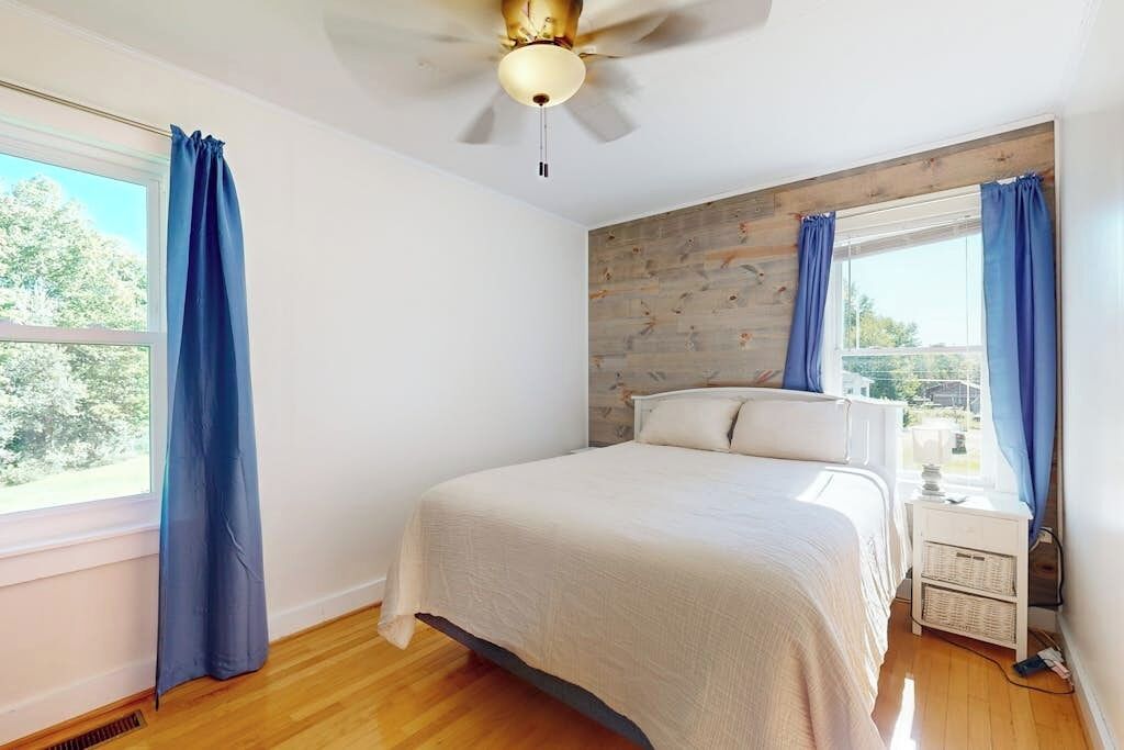 A bedroom with a bed, a ceiling fan, a window with curtains, and a wooden wall panel.