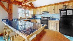 Une cuisine avec des poutres en bois, un réfrigérateur noir et un îlot central recouvert de carreaux, avec des tabourets.