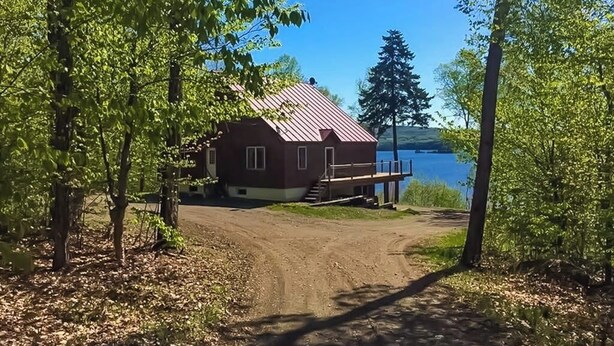 Une cabane avec un toit en métal, un chemin de terre menant à celle-ci, et un lac en arrière-plan.