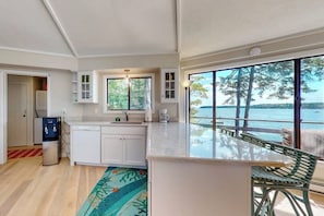 A kitchen with a view of the water, a dining area with a table and chairs, and a refrigerator. - Unique Oceanfront Home With a Deck, Tons of Natural Light, & Amazing Views (Wiscasset)